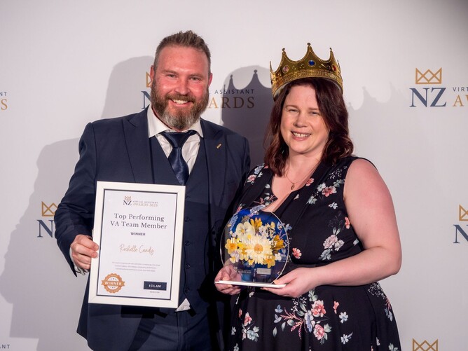 A man in a suit and a woman wearing a crown, both smiling and holding awards at the NZ Virtual Assistant Awards event, with a 'Top Performing VA Team Member' certificate and a floral trophy