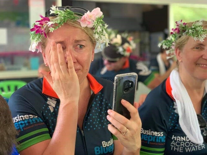 A woman wearing a flower crown, in a cycling jersey, wiping away a tear while holding her phone, beside her, another woman also wearing a flower crown
