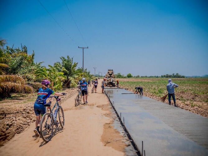 Cyclists walking beside freshly laid concrete on a road construction site in Thailand, with workers finishing the pavement under a clear blue sky