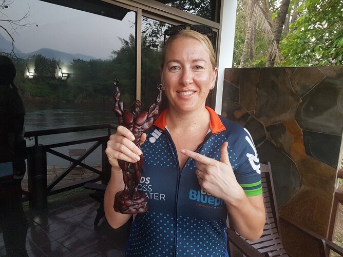 A woman in a cycling jersey holding with her right hand, a small statue of a figure posing in a bodybuilding pose, her left hand pointing at the statue while smiling
