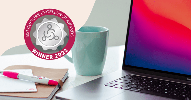 A close-up of a desk with a pink pen, notebook, and laptop, with a 'BSI Culture Excellence Awards Winner 2022' badge in the corner