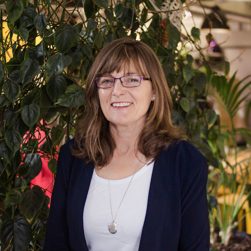 Smiling woman with glasses and shoulder-length brown hair, wearing a navy blazer and white top, standing in front of green plants