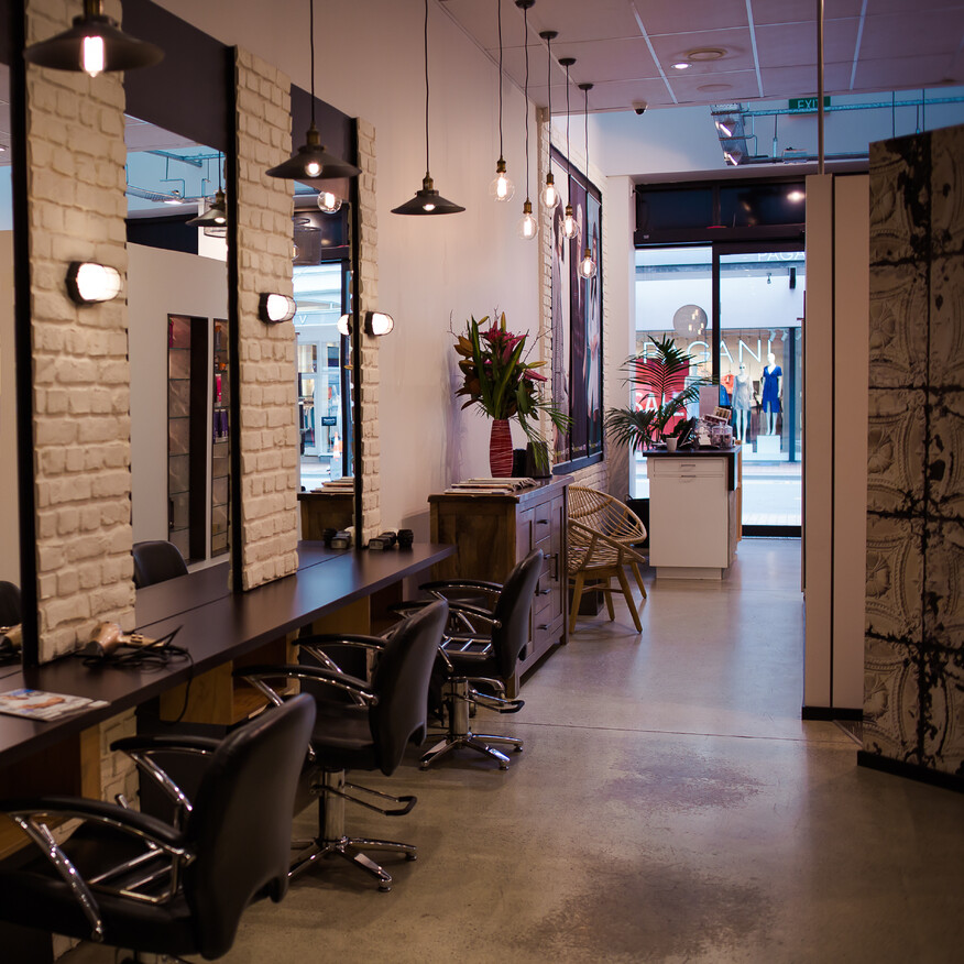 Modern hair salon with styling stations, large mirrors, black chairs, exposed brick walls, and pendant lighting, with a reception area in the background