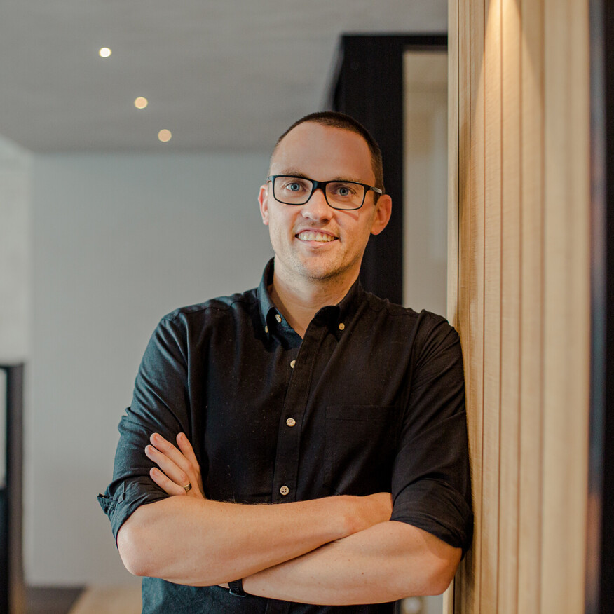 Smiling man with glasses wearing a black shirt, standing with arms crossed against a wooden panel in a modern office space