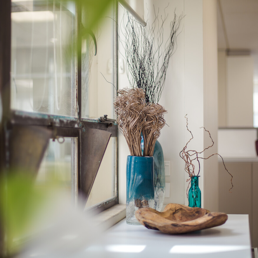 Bright office space with dried floral arrangements in blue glass vases and a wooden bowl on a white countertop near an open window