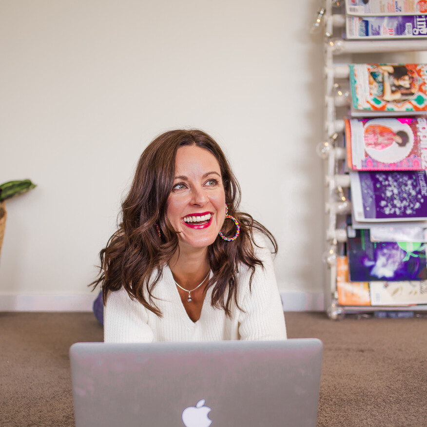 A smiling woman with wavy brown hair and bright red lipstick lies on the floor using a MacBook, with a magazine rack and woven basket in the background