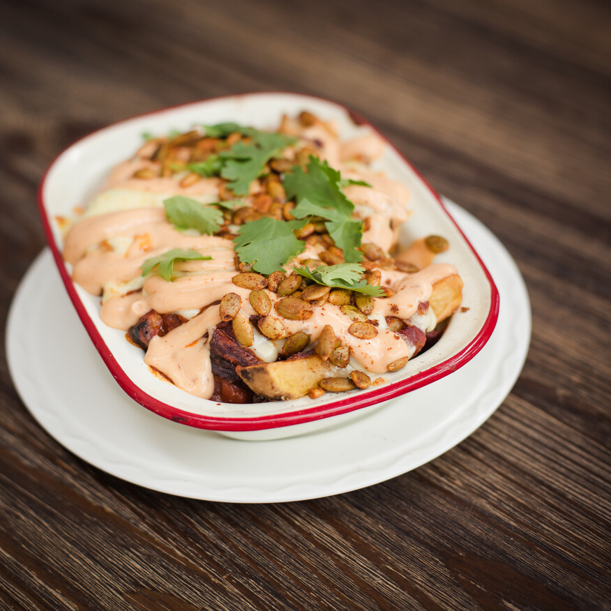 Enamel dish with loaded fries topped with creamy sauce, toasted seeds, fresh coriander, and spices, served on a wooden table