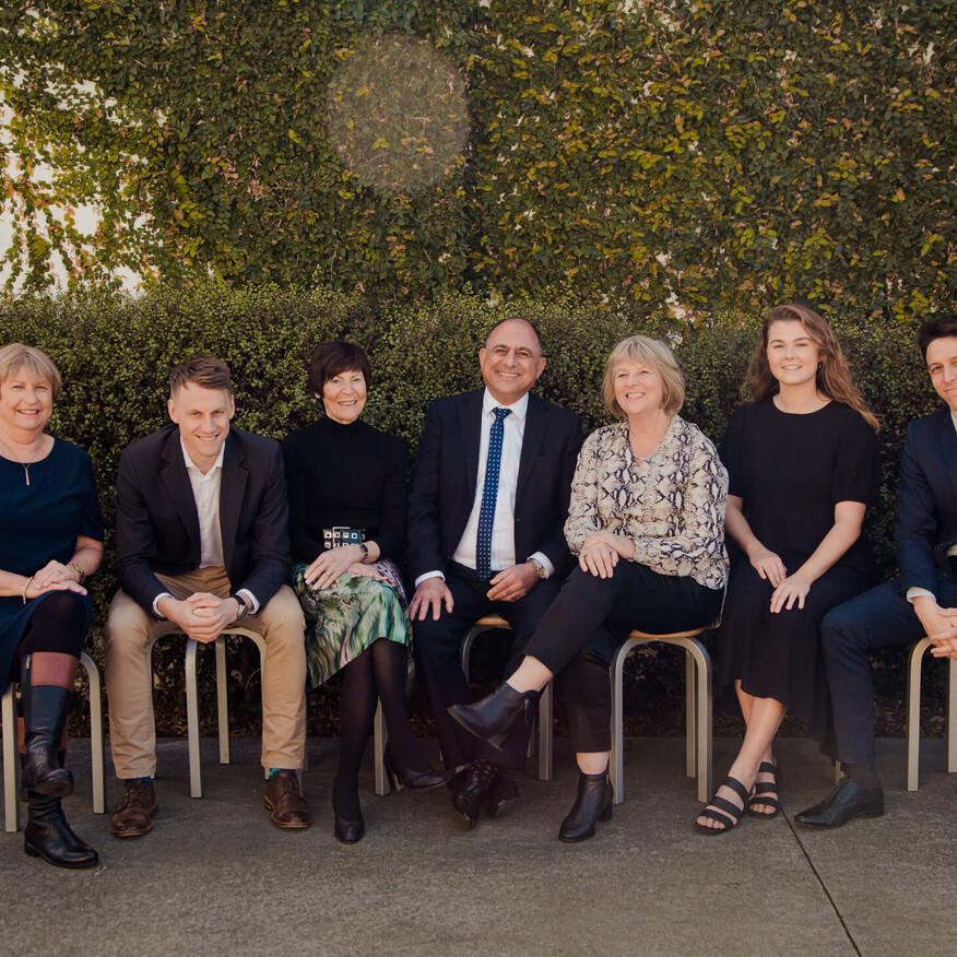 Seven professionally dressed people sitting on stools outdoors, smiling in front of a green hedge
