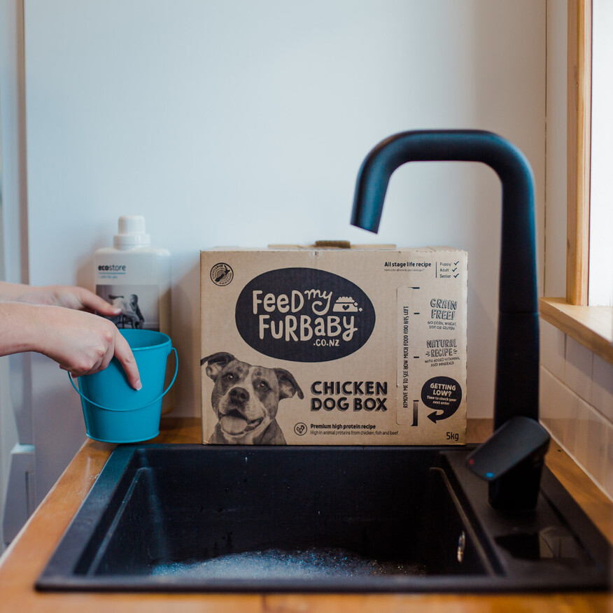 Person filling a blue bucket at a black kitchen sink, with a box of Feed My Furbaby chicken dog food on the counter