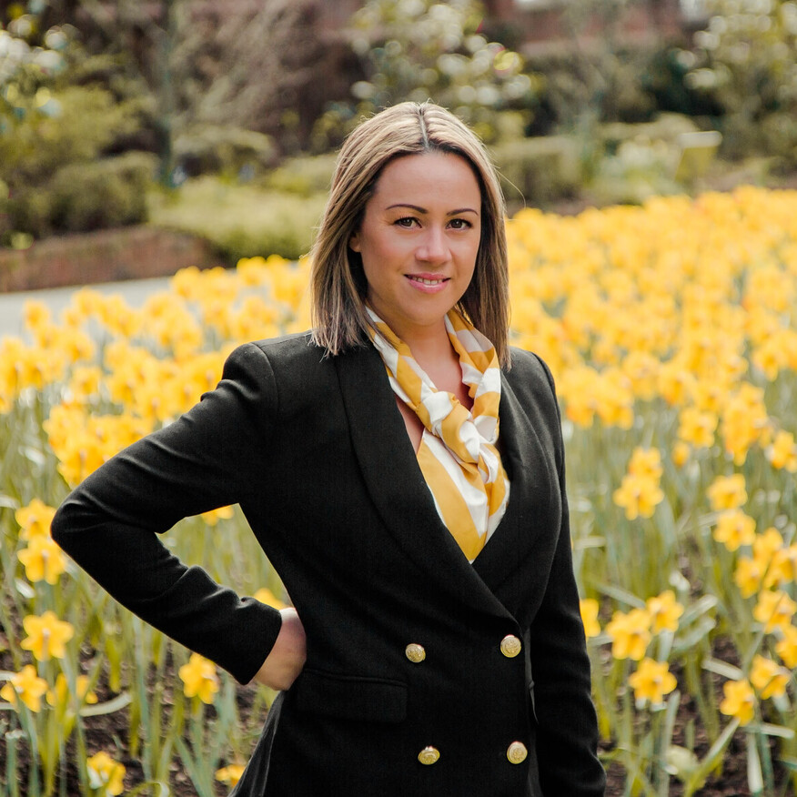 A woman in a black blazer and yellow scarf stands in front of a vibrant field of yellow daffodils, posing confidently with one hand on her hip