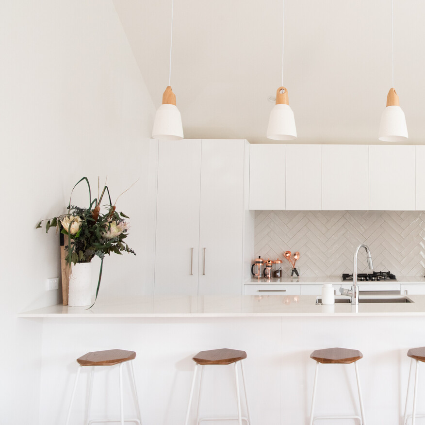 Modern white kitchen with a large island, wooden bar stools, pendant lights, and a vase of fresh flowers on the counter