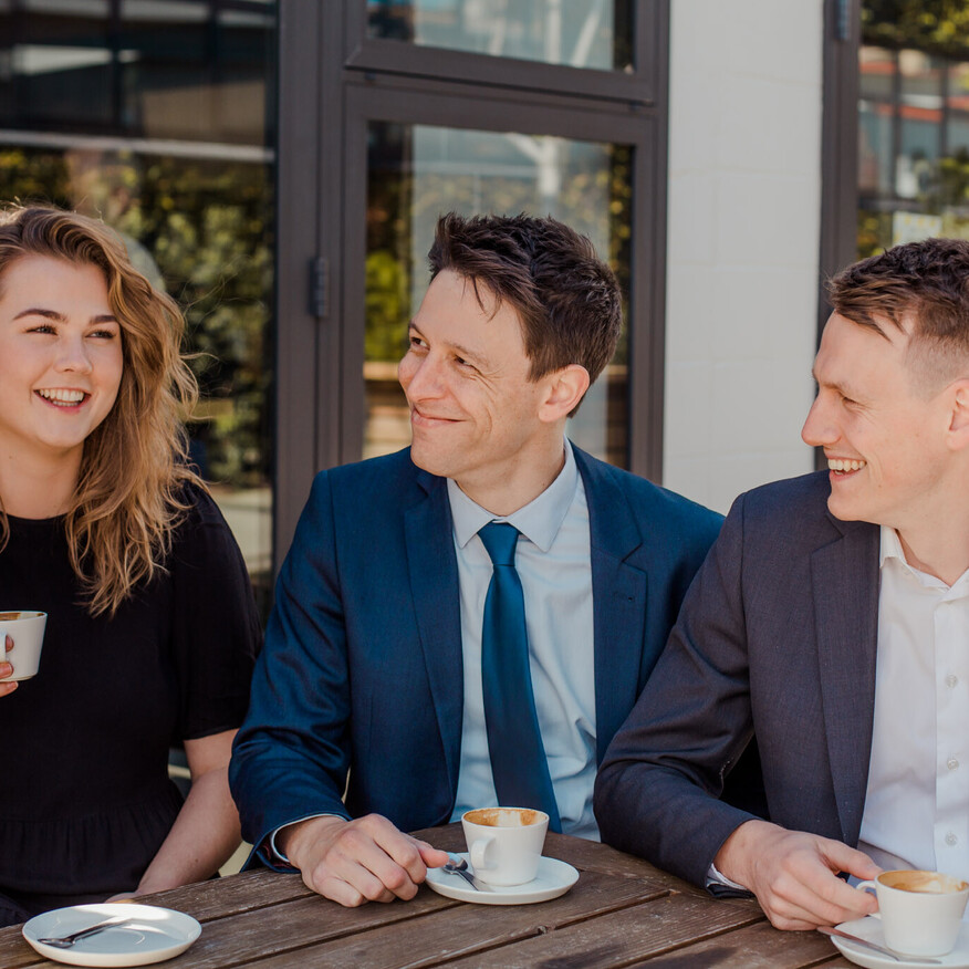 Three professionally dressed people sitting at an outdoor café table, smiling and chatting while holding cups of coffee