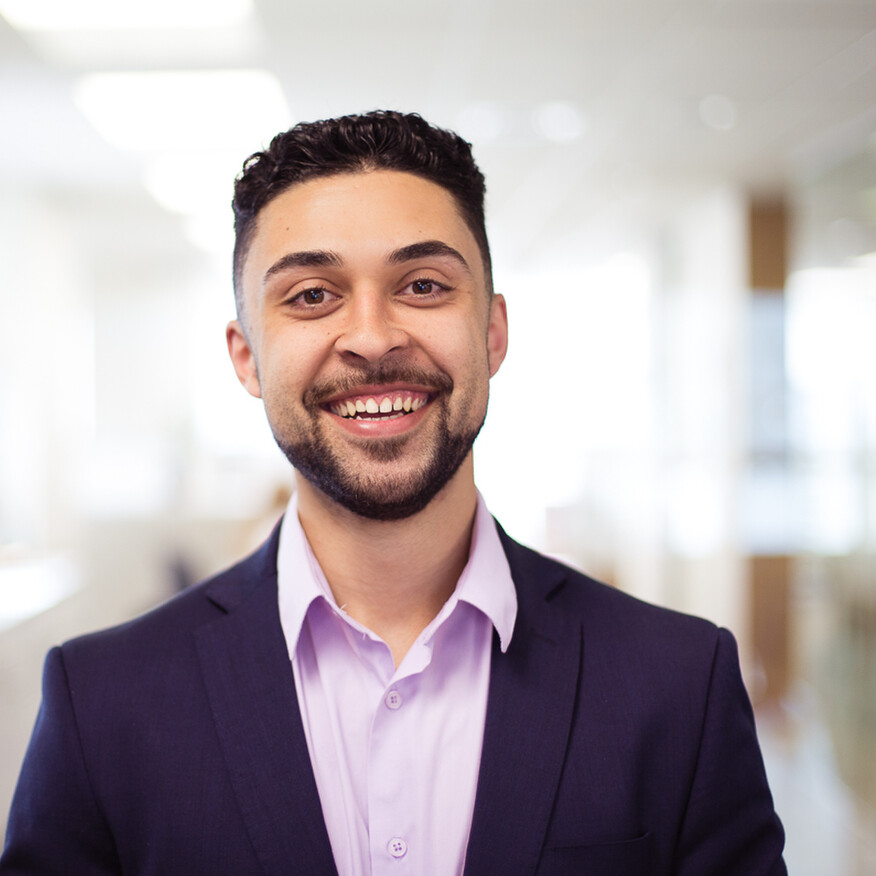 Smiling man wearing a suit and purple shirt, standing in an office environment with blurred background