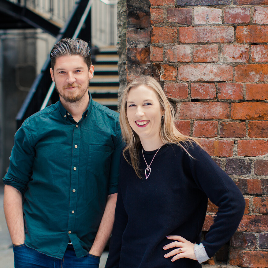 A man in a dark green button-up shirt and a woman in a black sweater with a heart pendant necklace smile while leaning against a rustic red brick wall in an urban setting