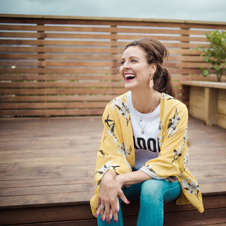 Woman sitting outdoors on a wooden deck, wearing a yellow floral kimono and a white t-shirt, smiling and laughing