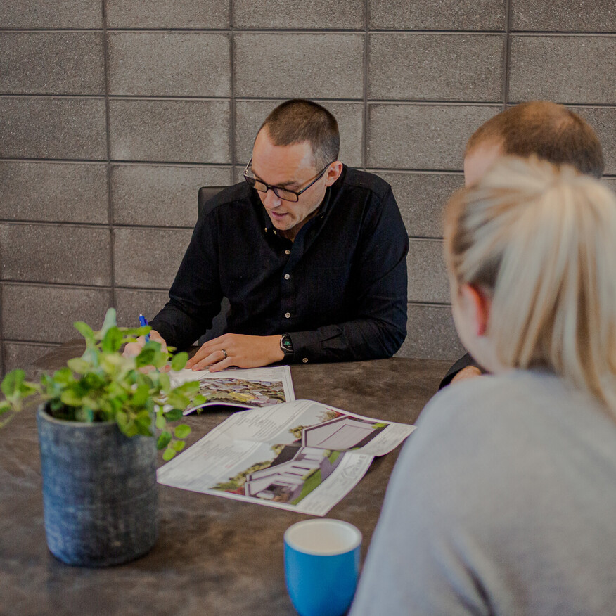 A group of three people in a meeting, reviewing architectural plans at a table with a plant and a blue cup visible