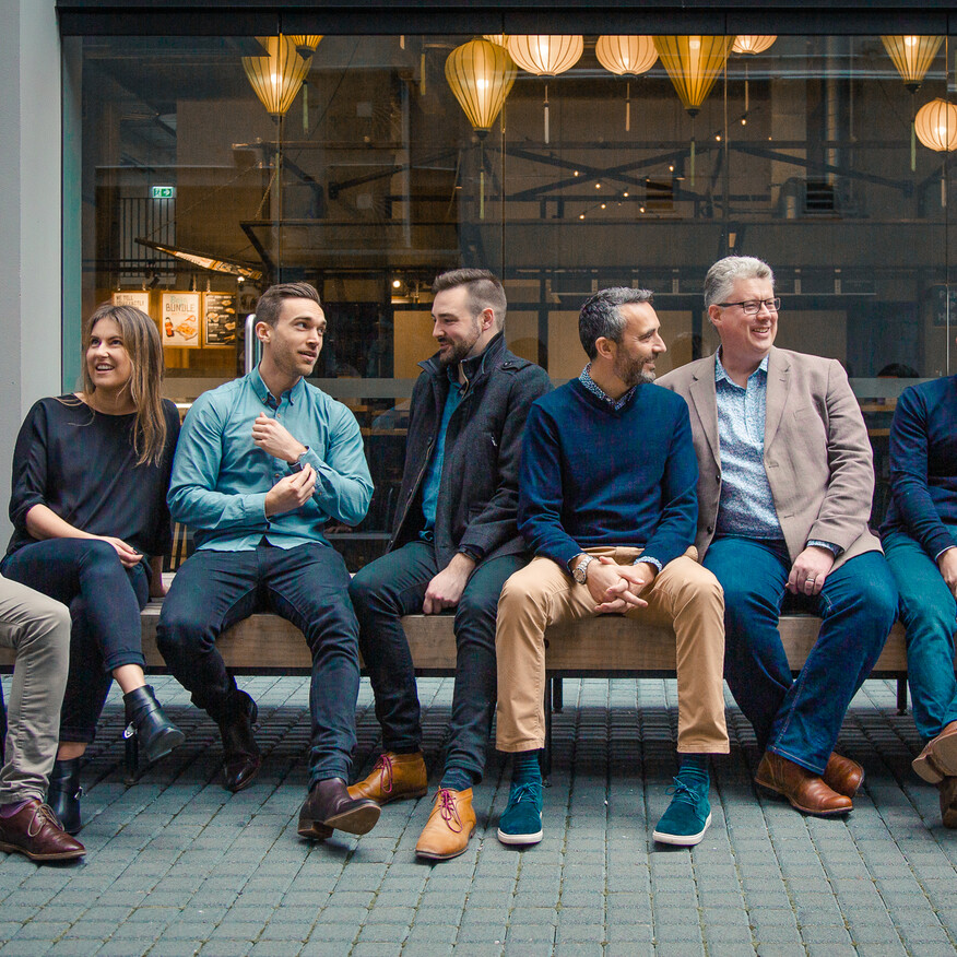 Group of nine casually dressed professionals sitting on a bench outside a modern café, engaged in conversation and laughter