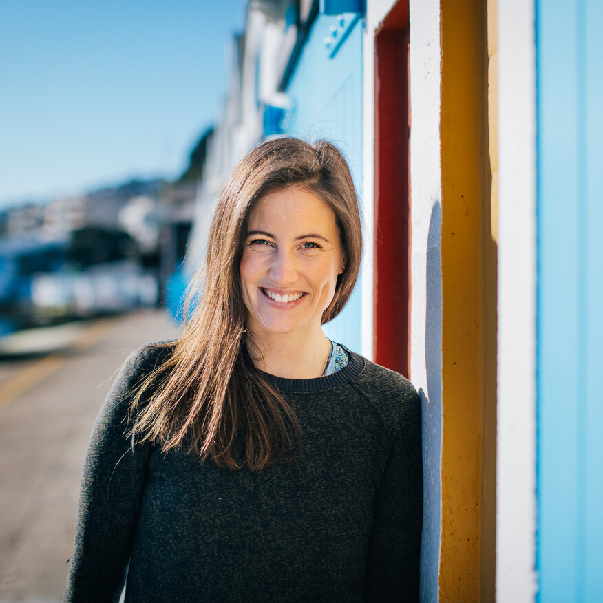 Smiling woman with long brown hair in a dark sweater leans against a colourful beach shed, with a marina and hillside buildings in the background