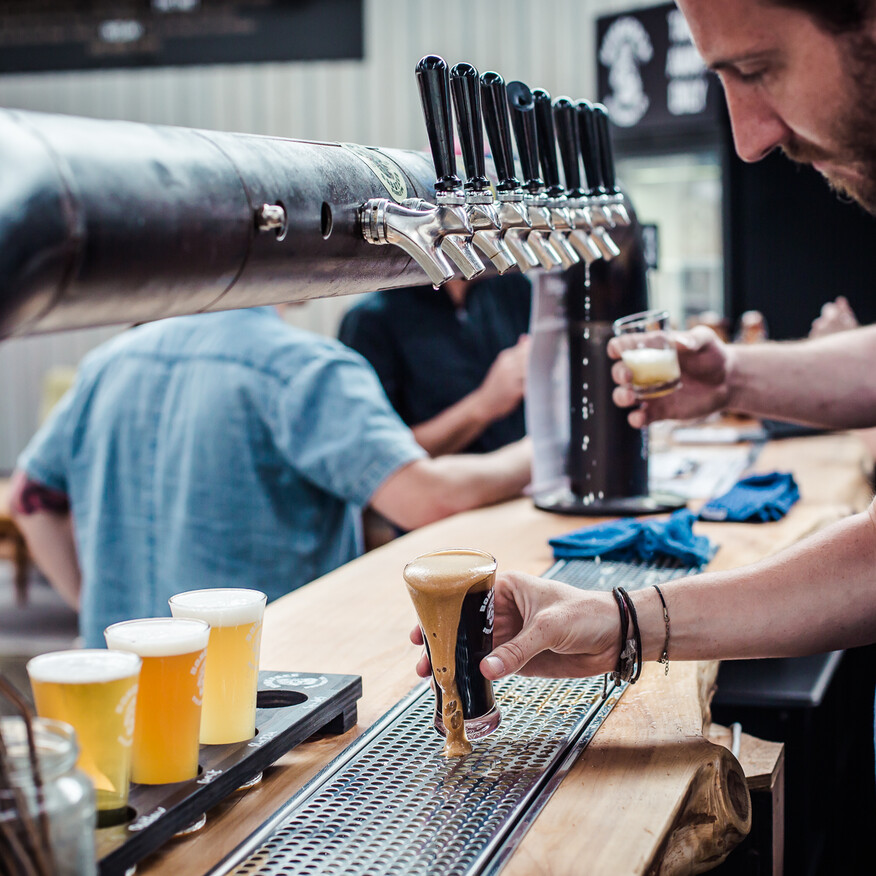 Bartender pouring a dark beer from a tap while holding a tasting glass, with a wooden bar and several pints of beer in the foreground