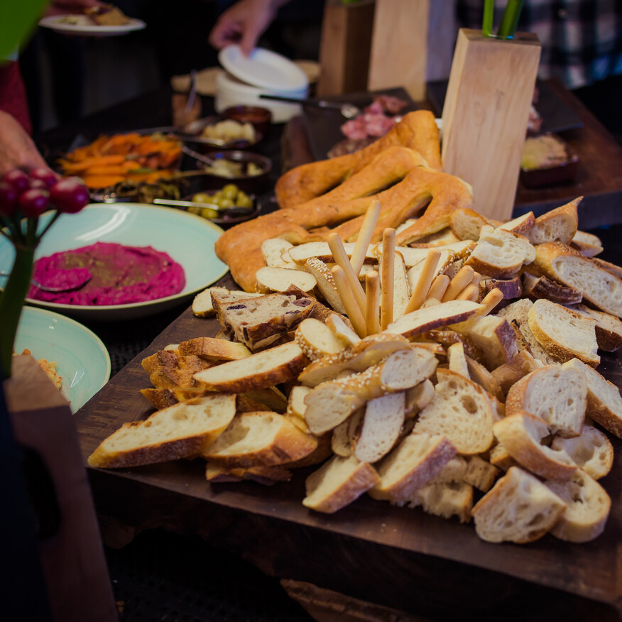 A rustic wooden platter filled with an assortment of sliced bread, breadsticks, and pretzels, surrounded by colorful dips, olives, and charcuterie at a buffet-style gathering