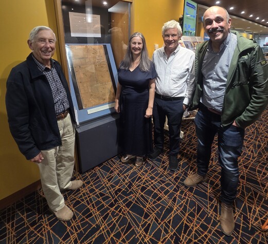 Tania and three men smile at the camera.  They are standing beside a case containing an early map of Auckland.