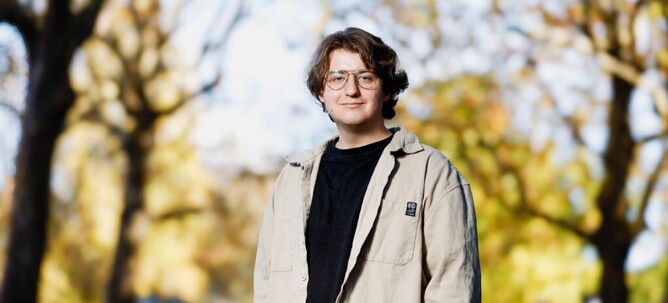 A young man is looking at the camera. He is wearing a t-shirt, jacket and glasses.