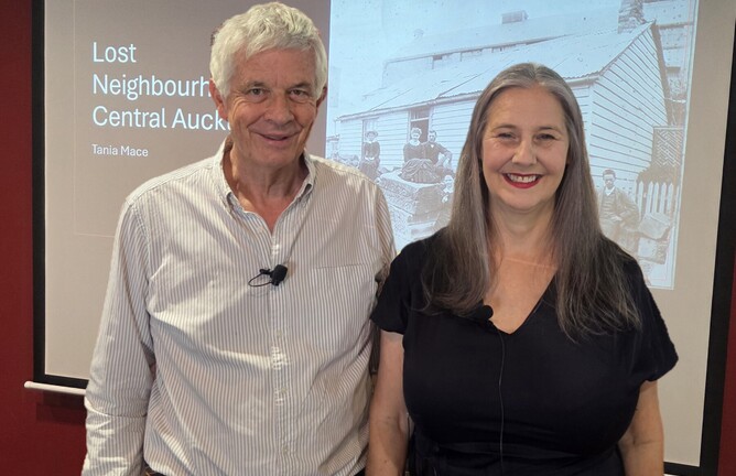 A man and a woman smiling at the camera. They are standing in front of a PowerPoint slide entitles 'Lost Neighbourhoods of Central Auckland. Tania Mace.' The slide shows a photo of an old house.