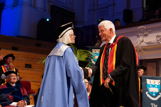 Graduation ceremony. People in academic regalia applaud as the Vice-Chancellor shakes David's hand. He is wearing a PhD gown and is smiling.