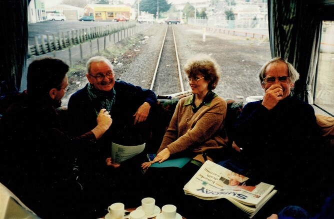 Three men and a woman are on a train; the tracks are visible through the window. They are smiling.