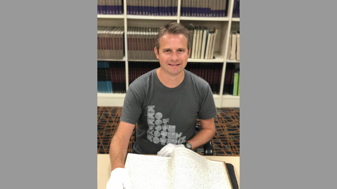 A man is in the Auckland Libraries heritage collections reading room. He is wearing gloves and looking at a manuscript. He is smiling at the camera.