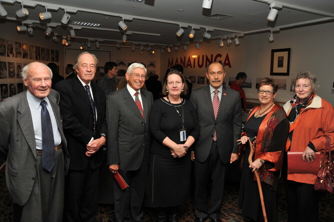 Group of four men and three women looking at the camera. One of the women is holding a carved stick. The Manatunga exhibition is visible in the background.