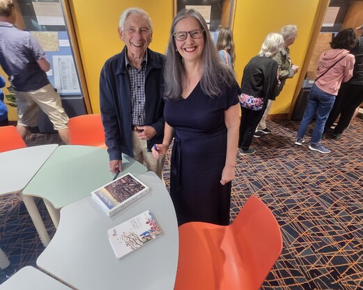 Tania and Colin Davis smile at the camera. Books and a pen are on a table in front of them.