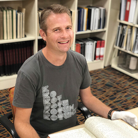 A man is in the Auckland Libraries heritage collections reading room. He is wearing gloves and a manuscript is open on the table in front of him. He is smiling at the camera.