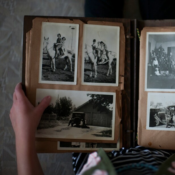 An old photo album resting on a persons lap with old photographs in it.