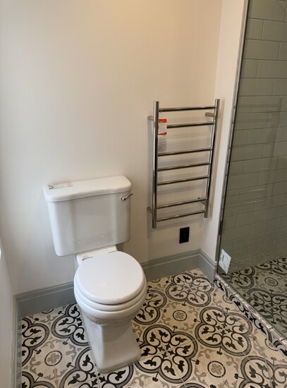 White burlington toilet in a bathroom with a black and white tiled floor, next to a chrome heated towel rail and glass shower