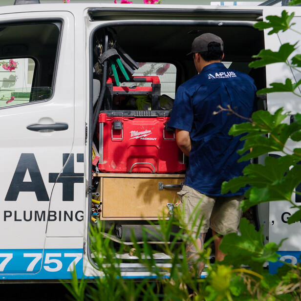 plumber with aplus plumbing and gas branding on the back of his shirt facing away from camera reaching into open door of branded van gathering tools for the job