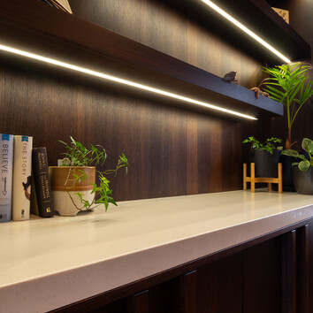 White desk with dark timber wall and built in shelving with lighting. 
