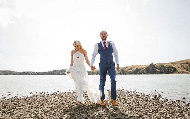 Photograph of bride and groom holding hadns walking on a rocky beach with water behind them using for a blog about not having a bridal party