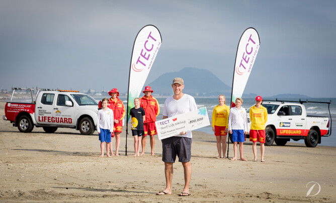 TECT General Manager Wayne Werder holds large $800,000 cheque on beach with lifeguards in background