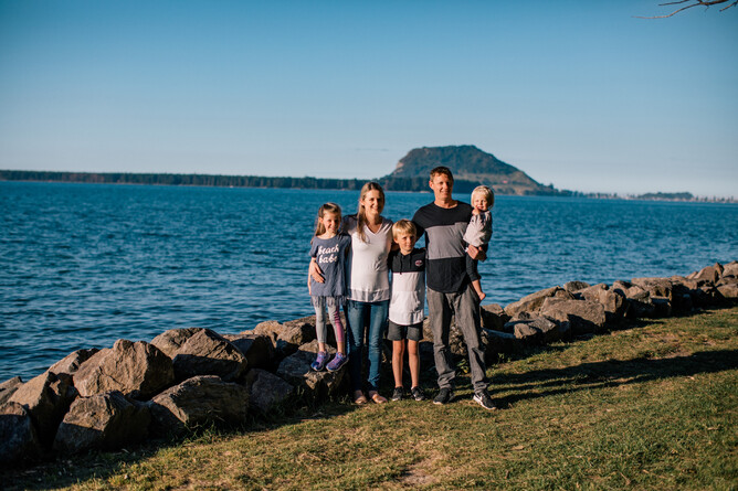 Parents Anton and Kayla stand with their three children in front of Mount Maunganui