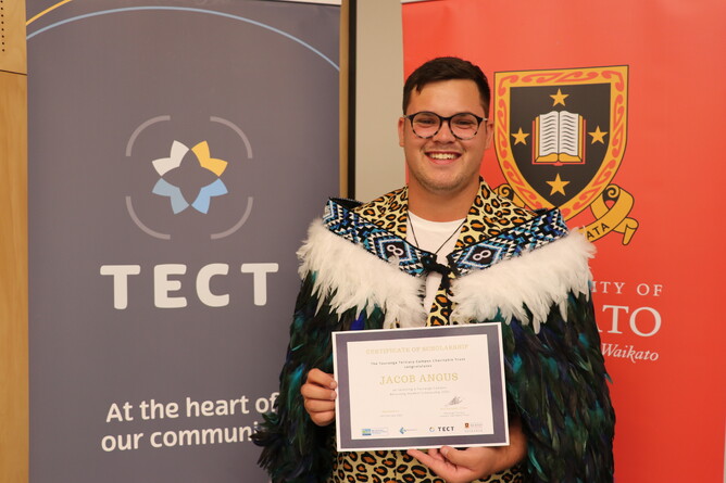 Successful scholarship recipient Jacob Angus poses with certificate of scholarship in front of TECT and University of Waikato banners