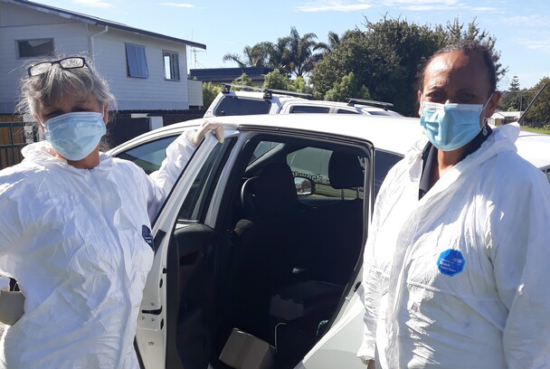 Two nurses in masks and PPE pose by car holding immunisation fridge