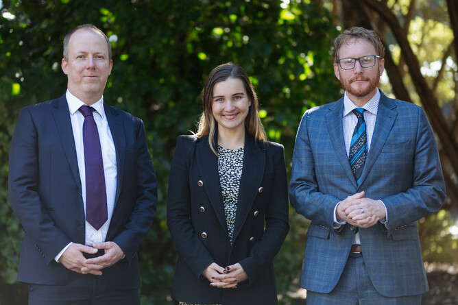A photograph of the three plaintiffs standing in front of a foliage background