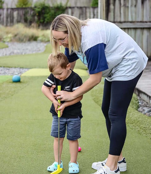 Mum and young son playing mini golf together, helping him hit the ball