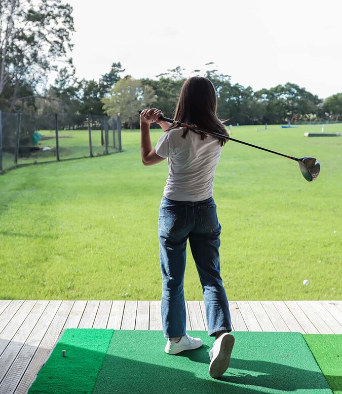 Young woman hitting golf club at driving range facing green