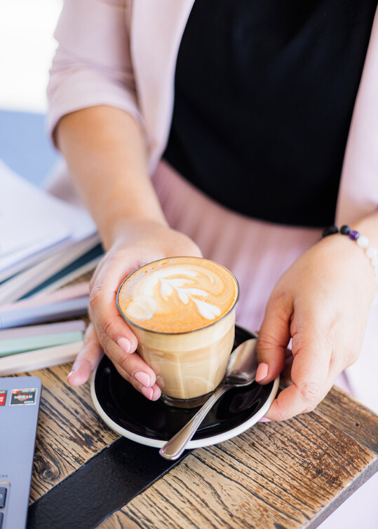 A woman holding a latte in a glass cup with latte art, seated at a wooden table with notebooks and a laptop - capturing a calm, focused moment during a CV consultation session.