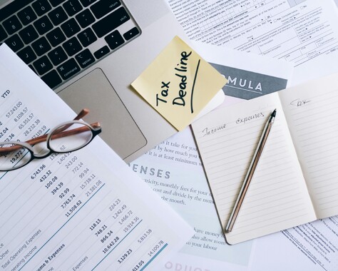 Laptop with Post-it note stating Tax Deadline and lots of paperwork lying around it, showing numbers and reports. Pencil and notepad.