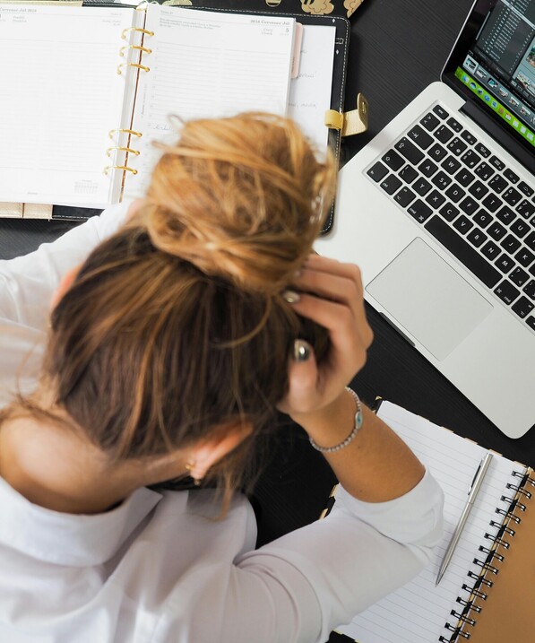 woman bending forward on her desk, drowning in paperwork, you can see her laptop, notebook / daily calendar and her hands holding up her head.