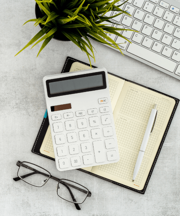 grey desk with a white keyboard, green pot plant, notebook with a pen, white calculator and glasses