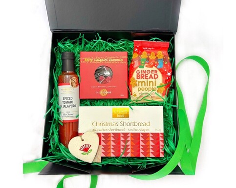 Christmas Treat Gift Box with Herb & Spice Hot Sauce, Sugarcrave Gummies, Bisko Gingerbread Mini People, Crave Shortbread, and Michelle Bow Ceramic Heart Ornament in black gift box with green filler.